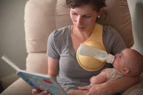 A woman is reading a book and simultaneously feeding her baby, who is cradled in her other arm. The baby drinks from a bottle supported by a hands-free device attached to the woman's shoulders, allowing for a comfortable and efficient feeding session.